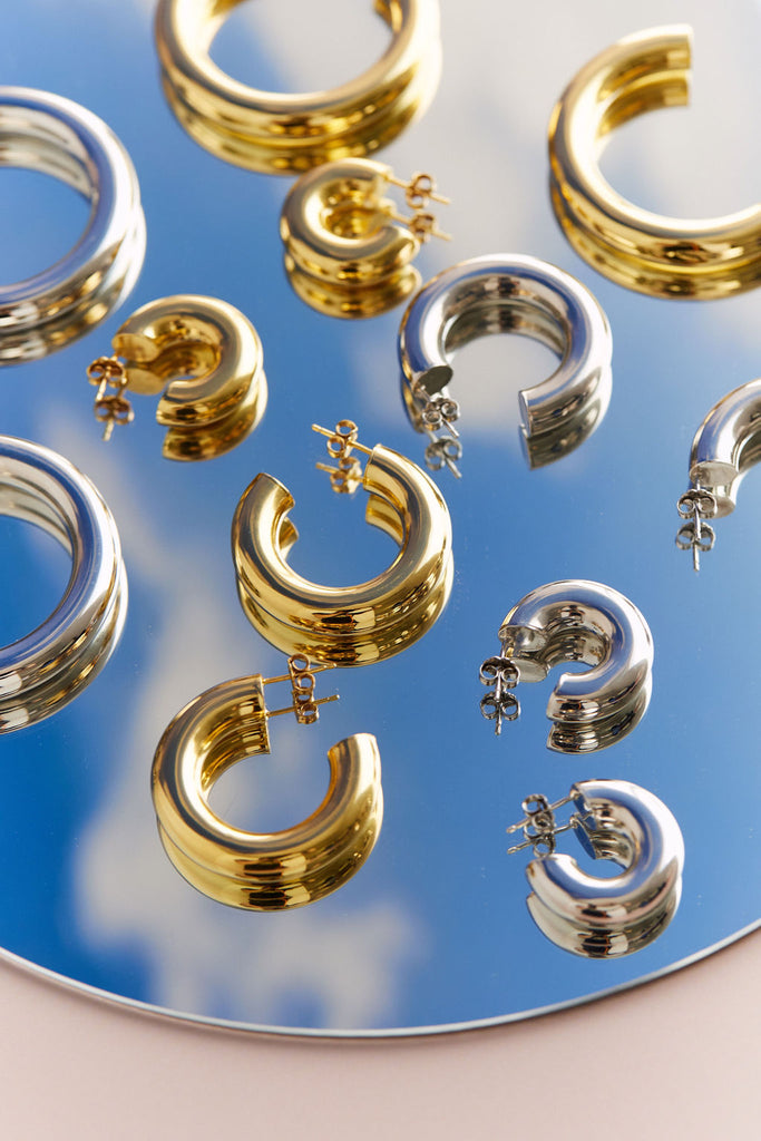 Styled product photography of the Ciambella Gold and Silver Huggie, Medium, and Large Hoops on a mirror with a sky reflection.
