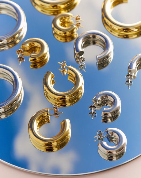 Styled product photography of the Ciambella Gold and Silver Huggie, Medium, and Large Hoops on a mirror with a sky reflection.